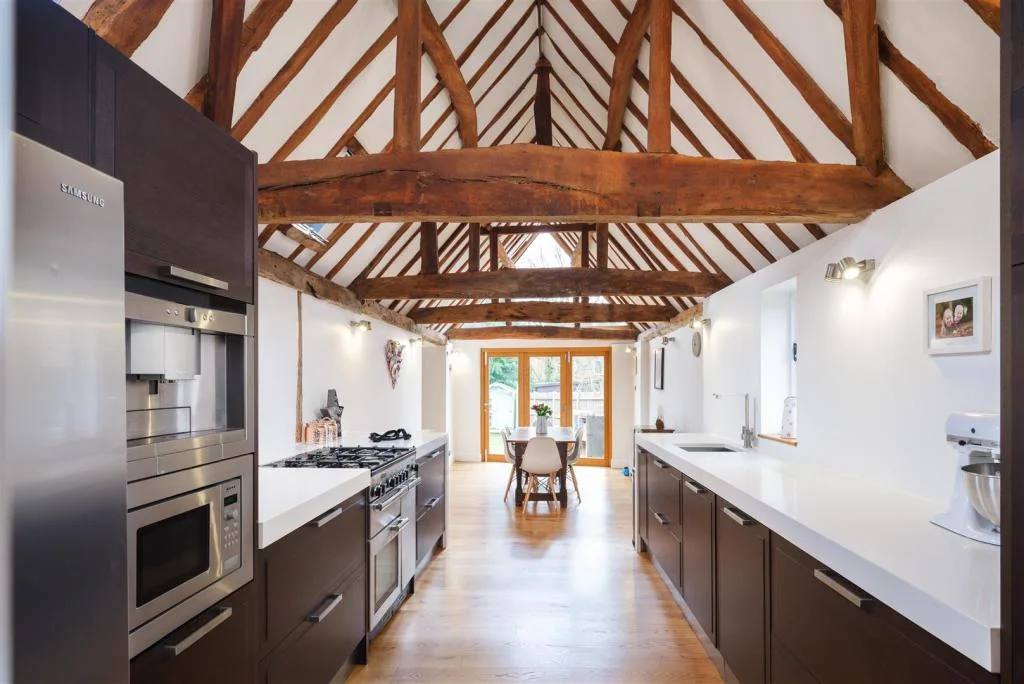Long galley kitchen with walnut units, steel bar handles and white waterfall-edge worktops beneath exposed oak roof beams, fitted by Jeremy Thornton